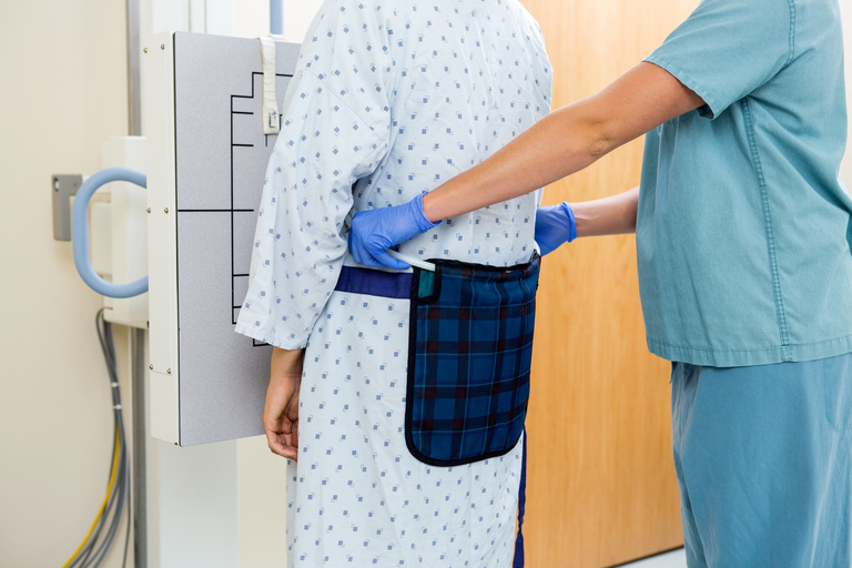 Midsection/cropped photo of nurse assisting patient standing in medical gown in front of z-ray machine.