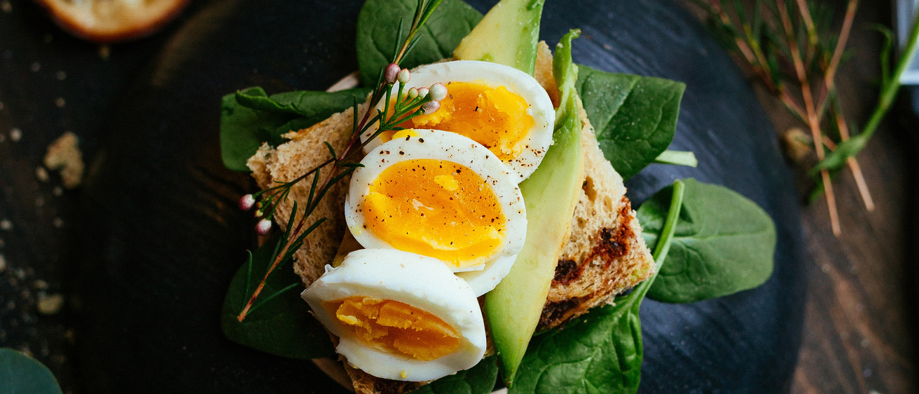 Black plate with spinach topped with toast, avocado, and sliced hard boiled eggs