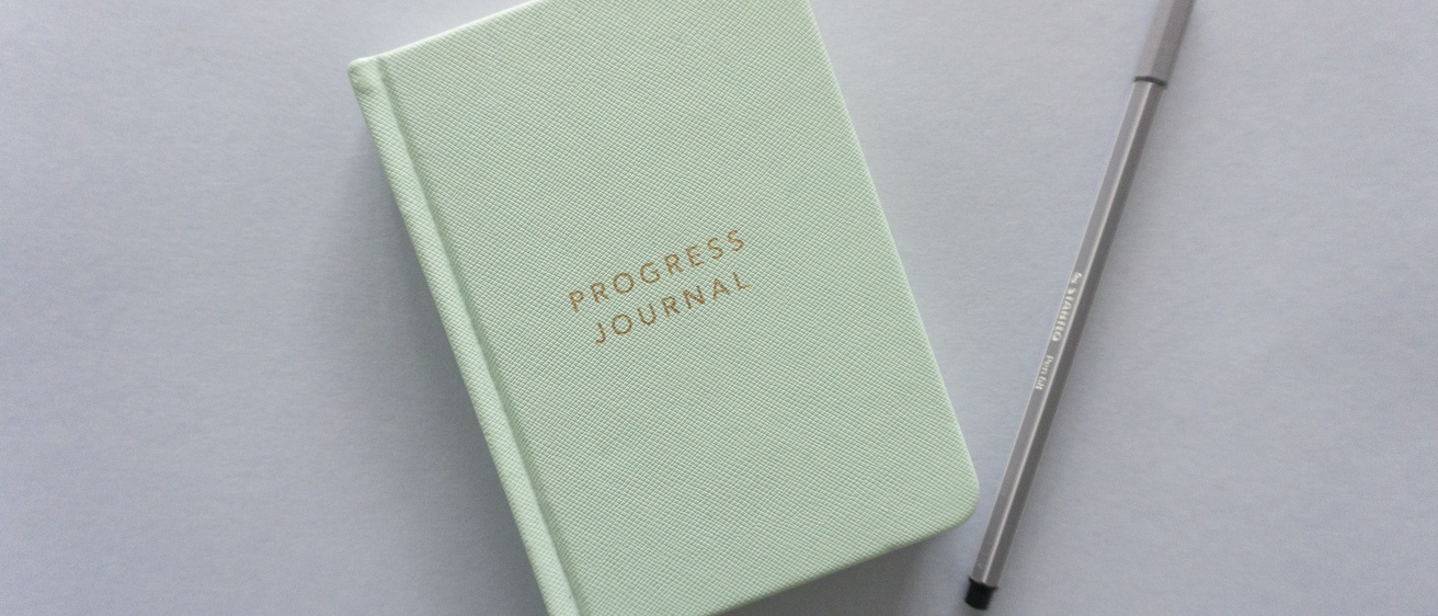 Light green journal with words "Progress Journal" on cover and pen laying on gray surface.