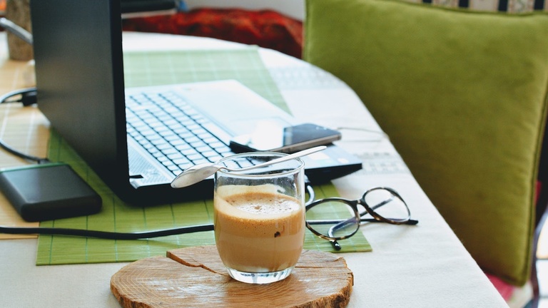 Glass of coffee, eyeglasses, and laptop computer on a table.