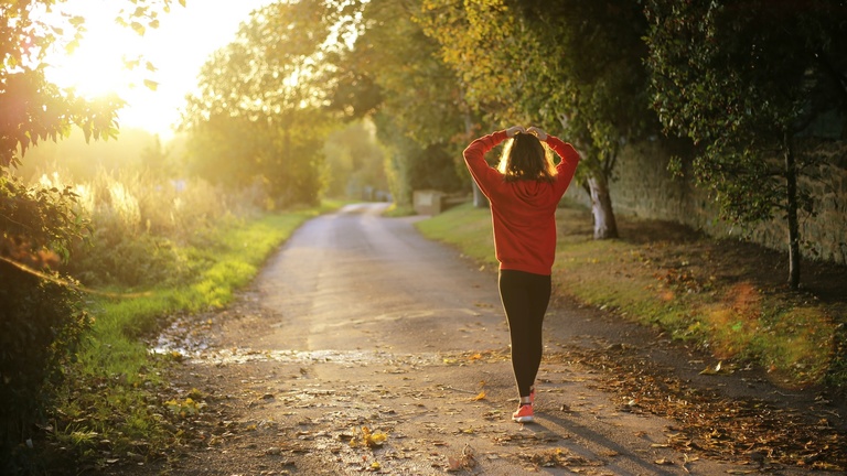 Person in workout clothes walking down wooded path toward a sunrise.